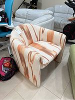 Front and left perspective of the barrel-style fabric chair with an orange and brown swirl pattern on beige background, showing cushioned seat and integrated armrests.