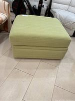 Front and top view of green fabric ottoman on wooden floor with nearby furniture and exercise equipment visible.