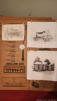 Three black and white ink art prints laid on a cardboard U-Haul box surface: a fishing boat art print, a cabin art print, and a smaller print showing a town street scene.