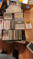 Overview of assortment of cassette tapes in cases arranged in a box, showing both clear and black cassette shells with labels and some with prints.
