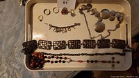 Overview of jewelry and accessories laid out on a tray: 5 rings, charm bracelet, multi-link fashion belt, silver-toned necklace with large oval textured discs, and beaded rosary with red and black beads.