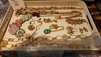 Wide view of the jewelry lot including multiple gold tone chains, floral brooches, rings, and earrings on white tray.