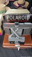 Polaroid Model J66 camera with original box and brown carrying case alongside hats in background.