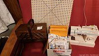 Wide view of the wooden sewing box closed and the cardboard box with sewing patterns, along with the sewing machine on a red chair.