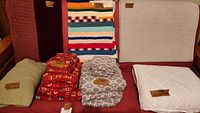 Three folded sheet sets (green, red with print, gray patterned) and three blankets (two knitted afghans and one white quilted cover) laid on a red couch.