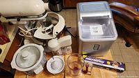 Four kitchen appliances arranged on floor and table: KitchenAid mixer, slow cooker, manual juicer, and ice maker.