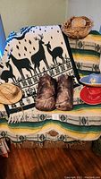 Photo showing 3 old west themed hats (2 straw hats, 1 blue wool hat with emblem), 1 adult wool poncho with stripes, 2 camo sleeping bags in bags, and 1 wool blanket with deer pattern.