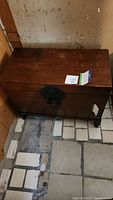 Full view of one vintage wooden trunk showing wood surface, metal latch, and legs on tiled floor.