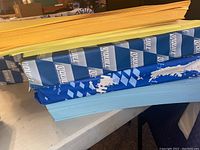 Stack of multiple paper reams in blue, yellow, and other colored packaging with loose paper on top