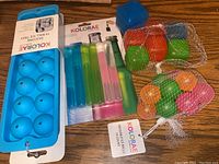 Blue silicone ice tray for spherical ice cubes, Kolorae brand, alongside reusable brightly colored ice cubes and drink chillers in tube shapes, shown on wooden surface.