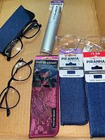 Photo of eyeglasses and cases on a cardboard background showing three fabric cases, four pairs of glasses including black, blue tinted, and clear framed glasses with labels and tags.