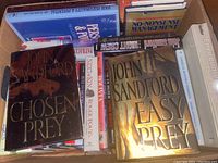 Top view of several books including 'Chosen Prey' and 'Easy Prey' by John Sandford, visible titles such as 'No-Nonsense Management', 'Environmental Geology', and others, arranged in a box.