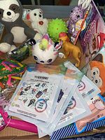Multiple plush toys including a sloth, unicorn, and white dog positioned around various kid's items such as over 10 packaged colorful kids face masks, a yellow horse toy, pom poms, wax lips candy, a pencil, and green spiky ball.