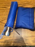 Closed blue umbrella with silver handle lying next to black mesh pouch containing rain poncho on a wooden surface.