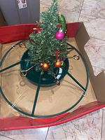 Green metal tree stand in box with a small artificial Christmas tree set inside it, decorated with colorful ornaments