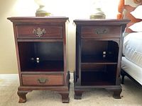 Two wooden bedside tables side by side showing full front view with drawer and lower compartment, featuring wear and ornate brass handles.