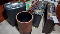 Overview photo of lot showing two briefcases, a paper shredder, and a decorative basket on carpeted floor next to furniture.