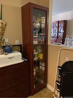Front view of entire tall display cabinet showing wood veneer finish and glass front door. Cabinet contains various canned goods and pantry items inside.