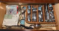 Overview of flatware and utensils organized in wicker compartments showing various forks, knives, serving spoons, decorative heart-shaped utensils and packaging visible.