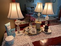Widely framed photo showing the pair of table lamps on a wood surface with a lace runner, mirror in background, vanity tray holding a Seth Thomas clock, jars, vase with flowers, candle and moisturizing cream set nearby.