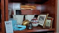 Shelf with two white Lenox vases, glazed pottery vase, British helmet souvenir bell, pewter dishes, framed pictures, and small boxes.