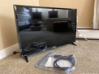 Full view of the LG television on carpet with remote and accessories in a plastic bag placed in front of it.