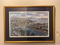 Framed lithograph with a scenic view of a waterfront town with houses and sailboats under a bright, partly cloudy sky.