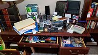 View of a wooden desk cluttered with office supplies, cordless phones, a receipt calculator, folders, pens, and VHS tapes.