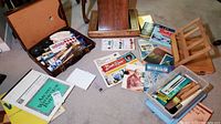 Full view of lot showing wooden art storage box, paint tubes, brushes, art books, watercolor pads, and easel on carpeted floor.