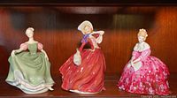 Three Royal Doulton figurines on wooden shelf: woman in green dress, woman in red dress with bonnet, woman in bright pink floral dress holding fan.