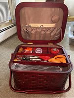 Open view of red wicker sewing basket showing interior with scissors, thread spools, pins, tape measure, and other sewing accessories organized in compartments.