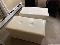 Pair of white vinyl upholstered storage benches shown from an angle on carpeted floor next to baseboard heater.