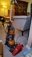 Dyson Ball upright vacuum with Dirt Devil handheld on carpet near chair and lamp in living room