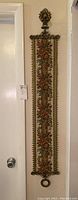 Full vertical view of the tapestry wall hanging revealing floral design, brass frame, hanging loops, and length compared to door.