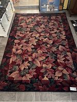Full view of the rectangular area rug laid out on tile flooring, showing the autumn leaf pattern in shades of red, green, orange, beige on a black background.