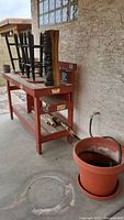 Angle view of red workbench with black bar stools on top, carved wooden totem, garden hose visible beside workbench, large terracotta planting pot in foreground.