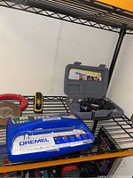 Shelf with Dremel hand drill set in gray case, 150-piece accessory kit in blue case, and measuring devices