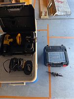 Overview of two drill sets laid out on table and ground: DeWalt 18V cordless drill kit with charger and batteries in black case; Black & Decker 14.4V Fire Storm drill kit with battery and drill bits in blue case.