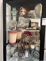 Glass display cabinet with various glass perfume bottles, gradient glass vases, decorative plates, shells, and porcelain mask on top shelf.