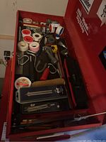 View of top toolbox drawer showing multiple rolls of Teflon tape, a pipe cutter, a spirit level, utility boxes, brushes, and hand tools mostly arranged flat.