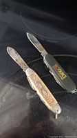 Photo showing two small pocket knives side by side with blades partially open on a dark surface.
