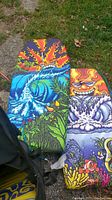 Three colorful boogie boards arranged lying on grass and stone, showing tropical wave and sun designs