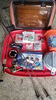 Photo showing the orange and brown plastic tackle box open with contents including fishing rod with reel, packs of bait and tackle, orange bucket with toys, and cooking sticks.