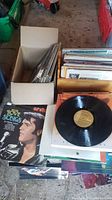 Two open cardboard boxes full of vinyl LPs stacked on floor; Elvis Love Songs cover and loose black vinyl disc visible