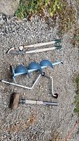 Manual ice auger with blue blade, large hedge clippers, and sledgehammer all placed on gravel ground.