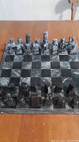 Full view of chessboard with all pieces set up in starting positions, showing black and white marbled squares and stone pieces with varying geometric carved shapes.