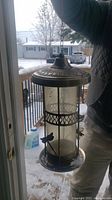 Full view of metal bird feeder showing cylindrical glass container and hanging hook, outdoor winter background