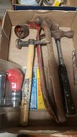 Box with three hammers visible: one claw hammer with light wooden handle, one hammer with red chipped paint handle, one ball peen hammer with black metal handle. Also visible is a metal pry bar and part of a boxed drill attachment.