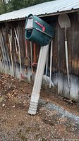 Rubbermaid mailbox and post leaning against a barn wall outdoors with garden tools hung in the background