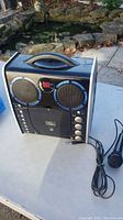 Front angle view of the portable karaoke machine on a white table outdoors, showing dual speakers with blue rings, cassette player, control buttons and knobs, carrying handle on top, and attached wired microphone coiled beside it.